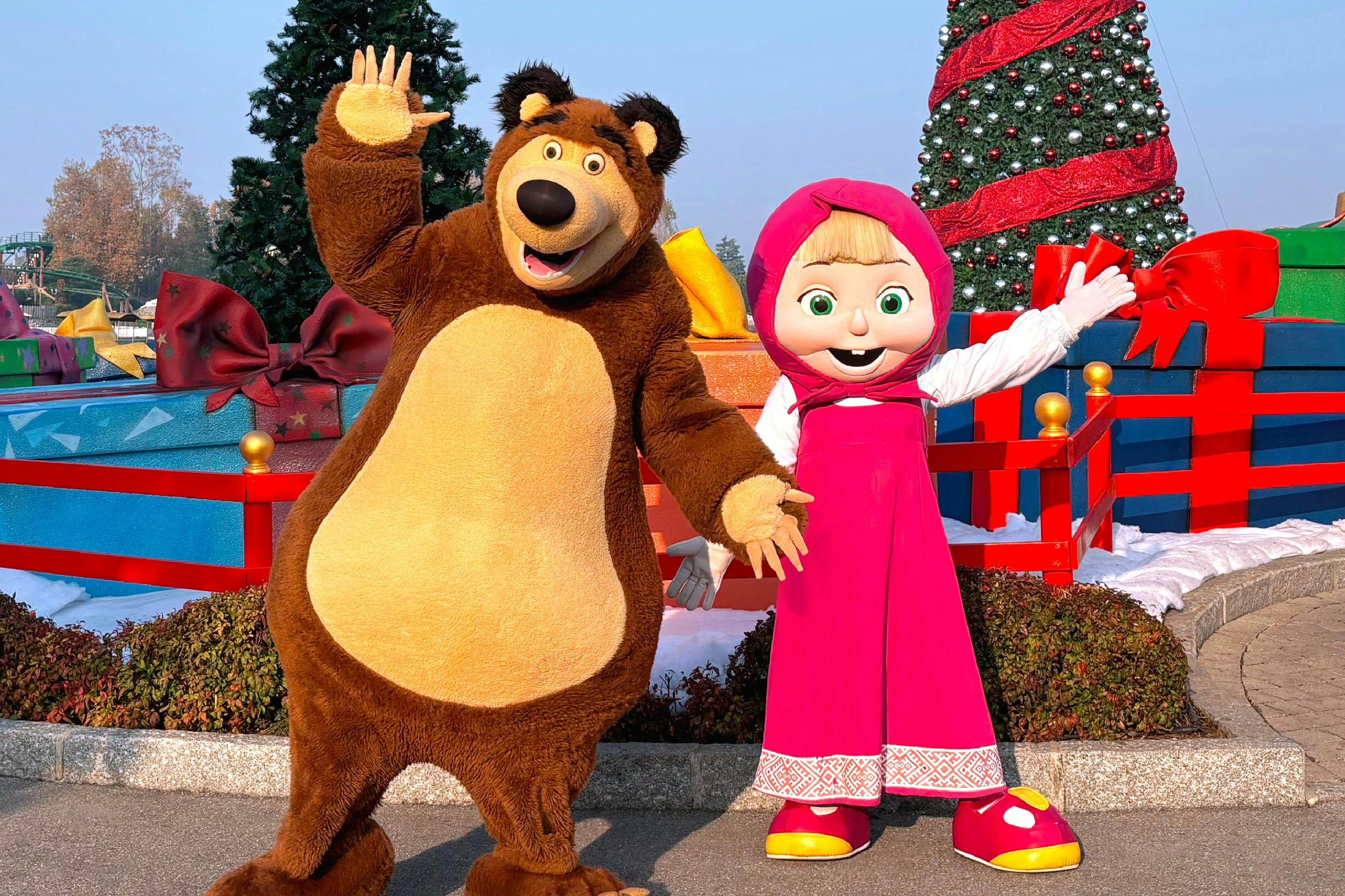 Δύο μασκοφόροι χαρακτήρες, μια αρκούδα και ένα κορίτσι, ποζάρουν μπροστά από ένα στολισμένο χριστουγεννιάτικο δέντρο και μεγάλα κουτιά δώρων.