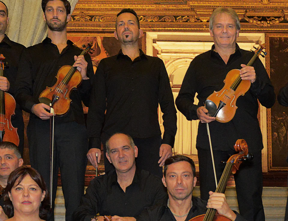 A group of musicians holding string instruments poses on an ornate staircase with detailed gold decorations in the background.