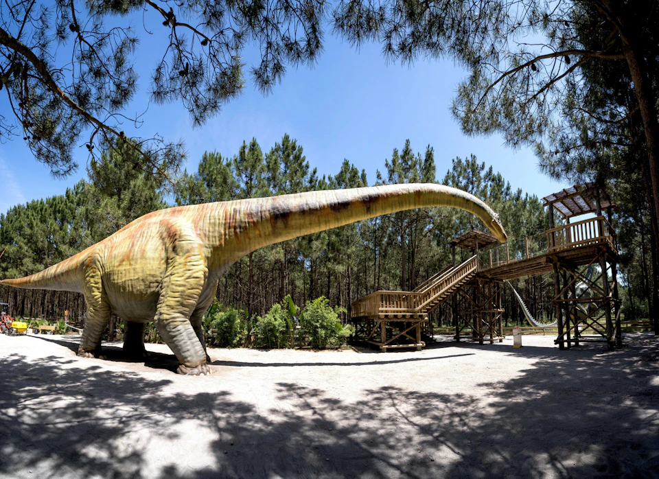 A giant dinosaur statue near a wooden structure set amidst a forest of tall pine trees under a clear blue sky.