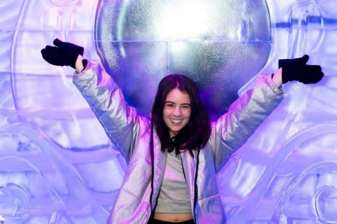 Une personne souriant et posant les bras levés, vêtue d'une veste réfléchissante, sur un fond lumineux violet et bleu.