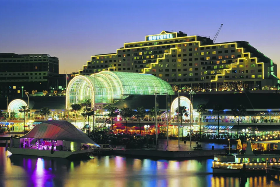 An illuminated urban waterfront scene at dusk featuring a glass-domed building, colorful lights, and reflections on the water.