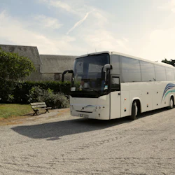 En parkerad vit turnébuss på en grusplats nära en byggnad med en träbänk och grönska i bakgrunden.