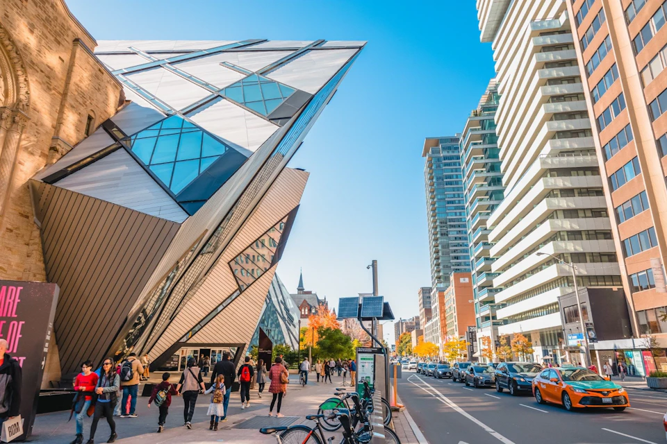 A modern, angular building beside a street with pedestrians, parked bicycles, and cars under a clear blue sky.