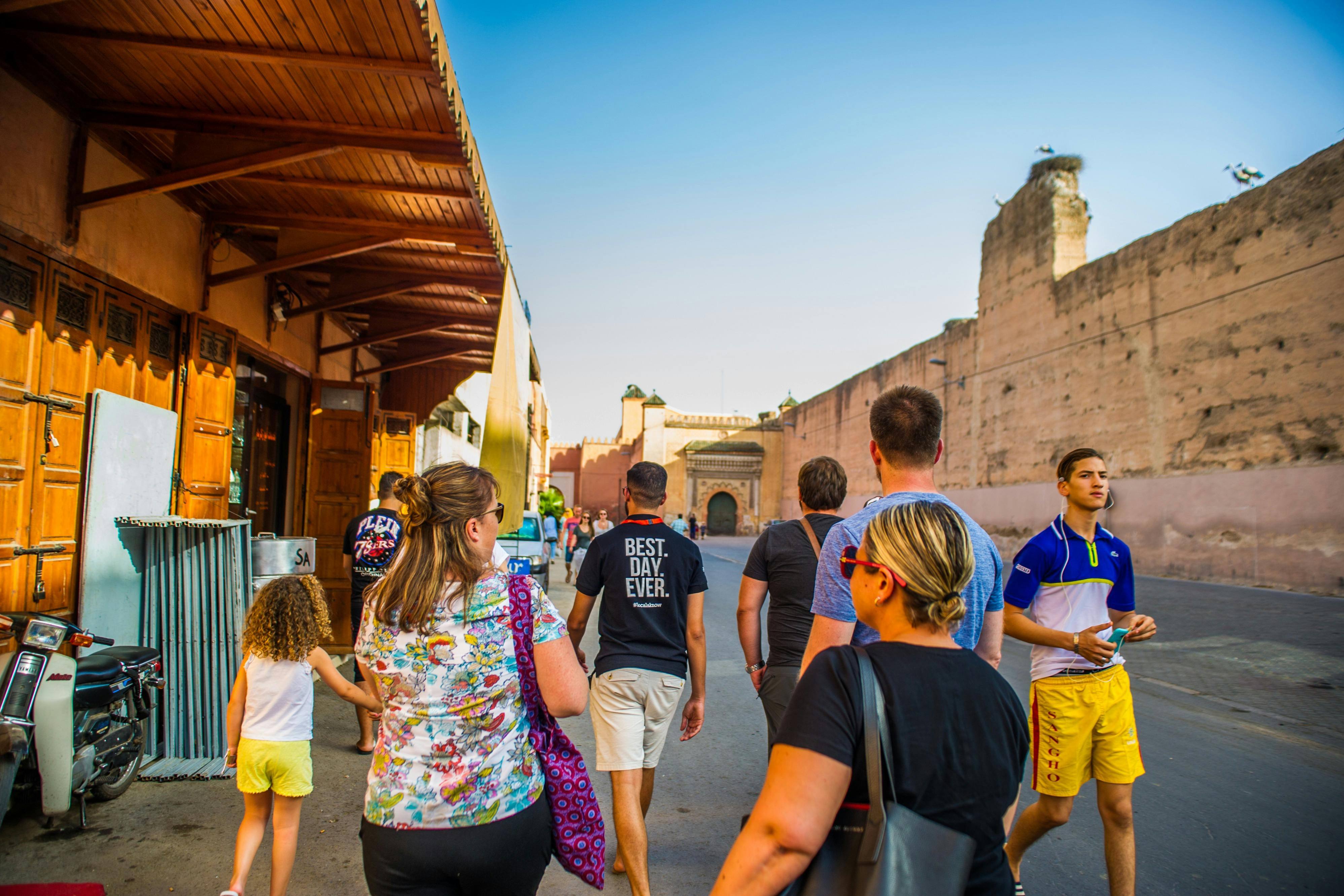 People walking along a street beside a large ancient wall under a clear sky. One person wears a shirt that says "Best. Day. Ever."