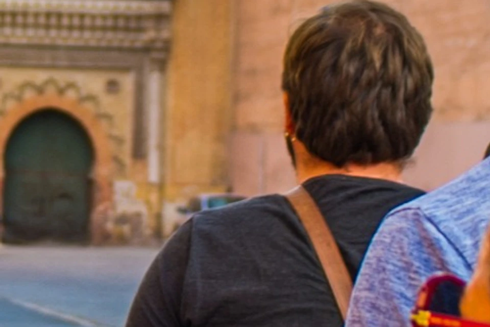 People walking along a street beside a large ancient wall under a clear sky. One person wears a shirt that says "Best. Day. Ever."