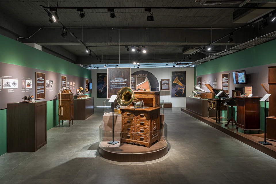 A museum exhibit featuring vintage gramophones and cabinets, displayed in a well-lit room with informational panels on the walls.