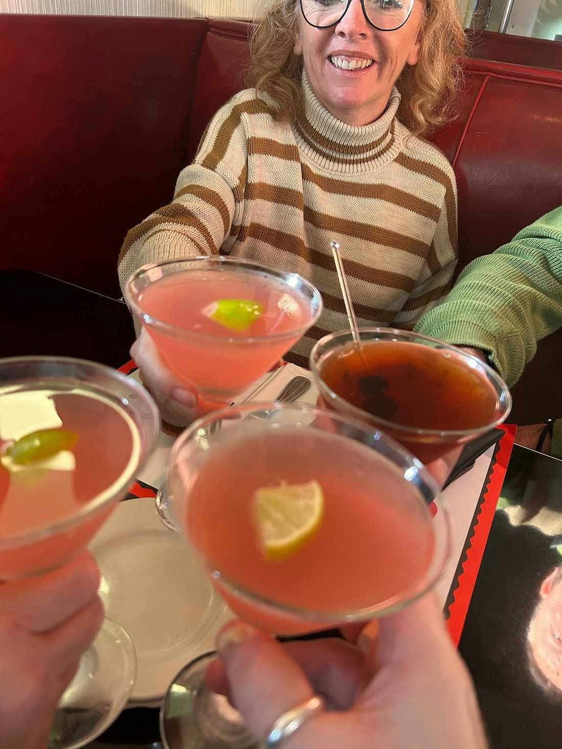 Person in a striped sweater holding a cocktail glass with lime, sitting in a red booth with three other drinks on the table.