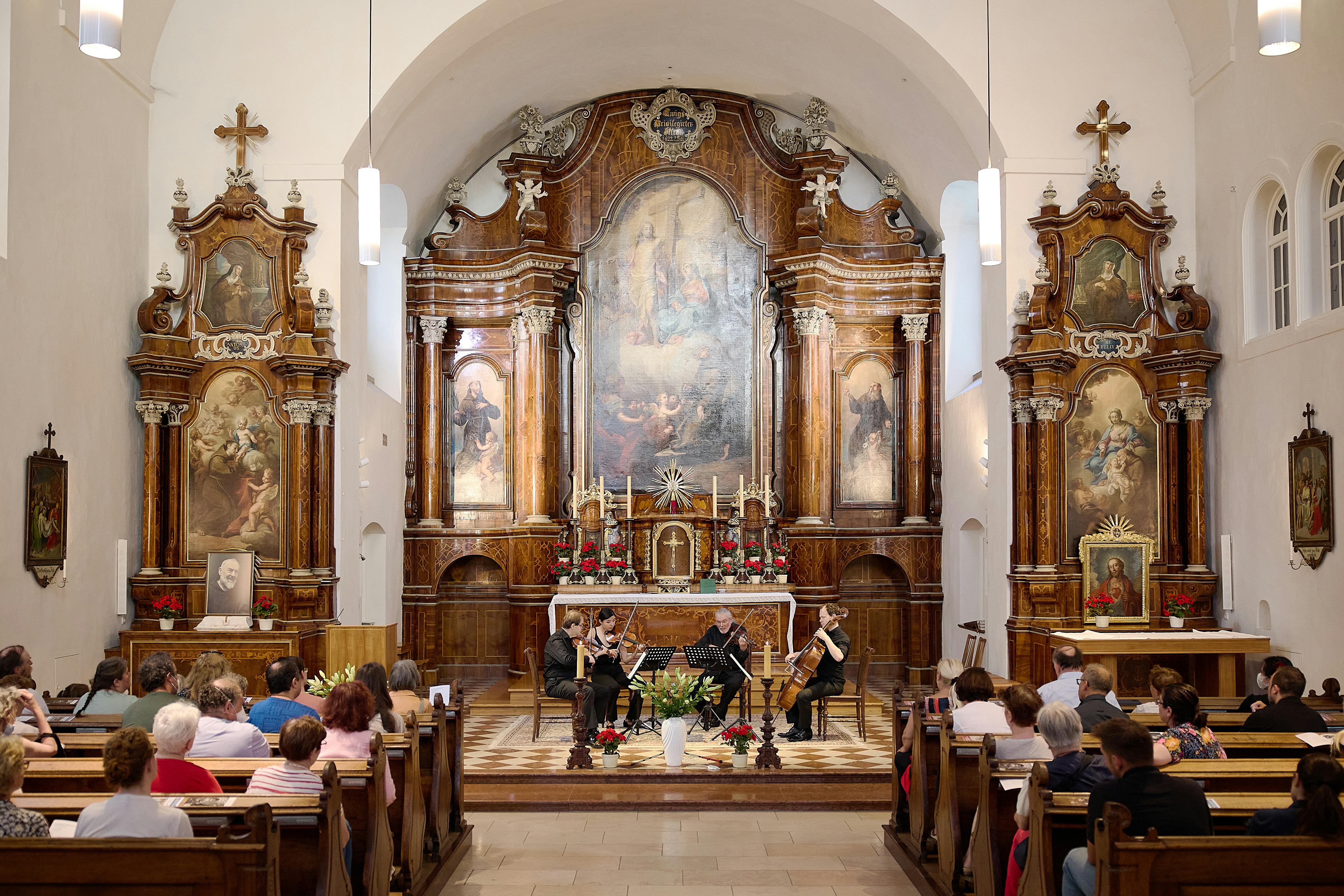 En stråkkvartett uppträder i en kyrka med människor sittande i kyrkbänkar, under utsmyckade religiösa konstverk och dekorationer.
