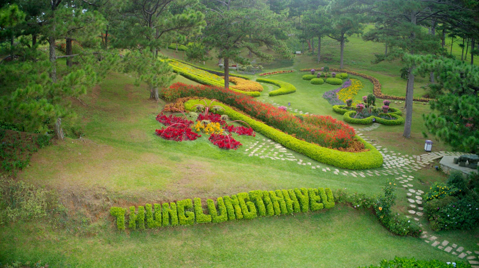 A lush garden with colorful flower beds, well-trimmed hedges, and trees, featuring "THUNG LUNG TINH YEU" shaped in greenery.