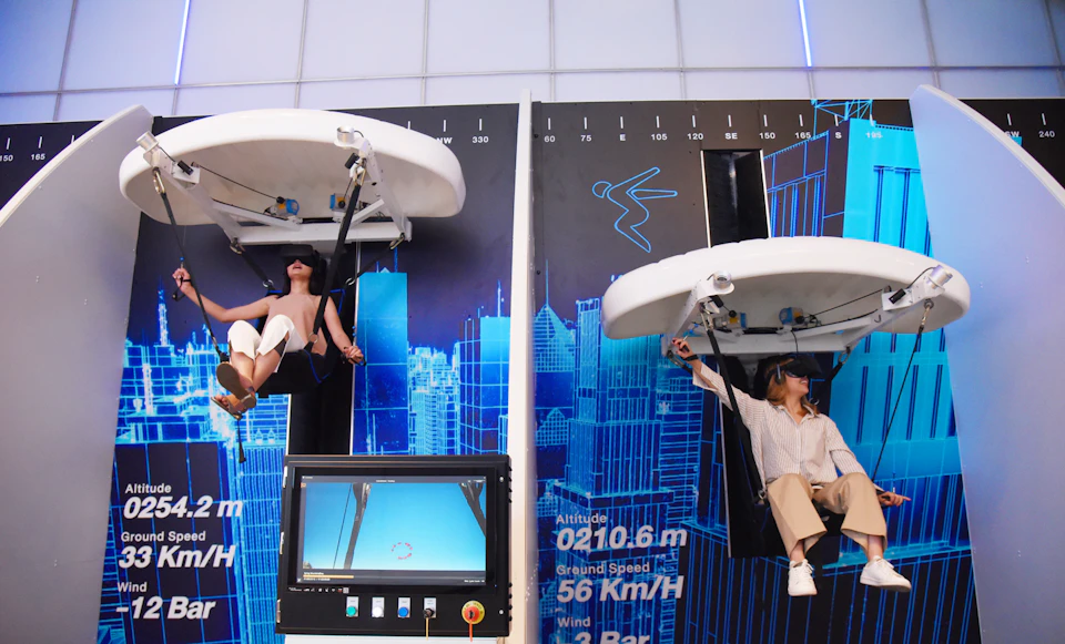 Two people sit in virtual reality hang gliding simulators against a blue cityscape backdrop with metrics displayed.