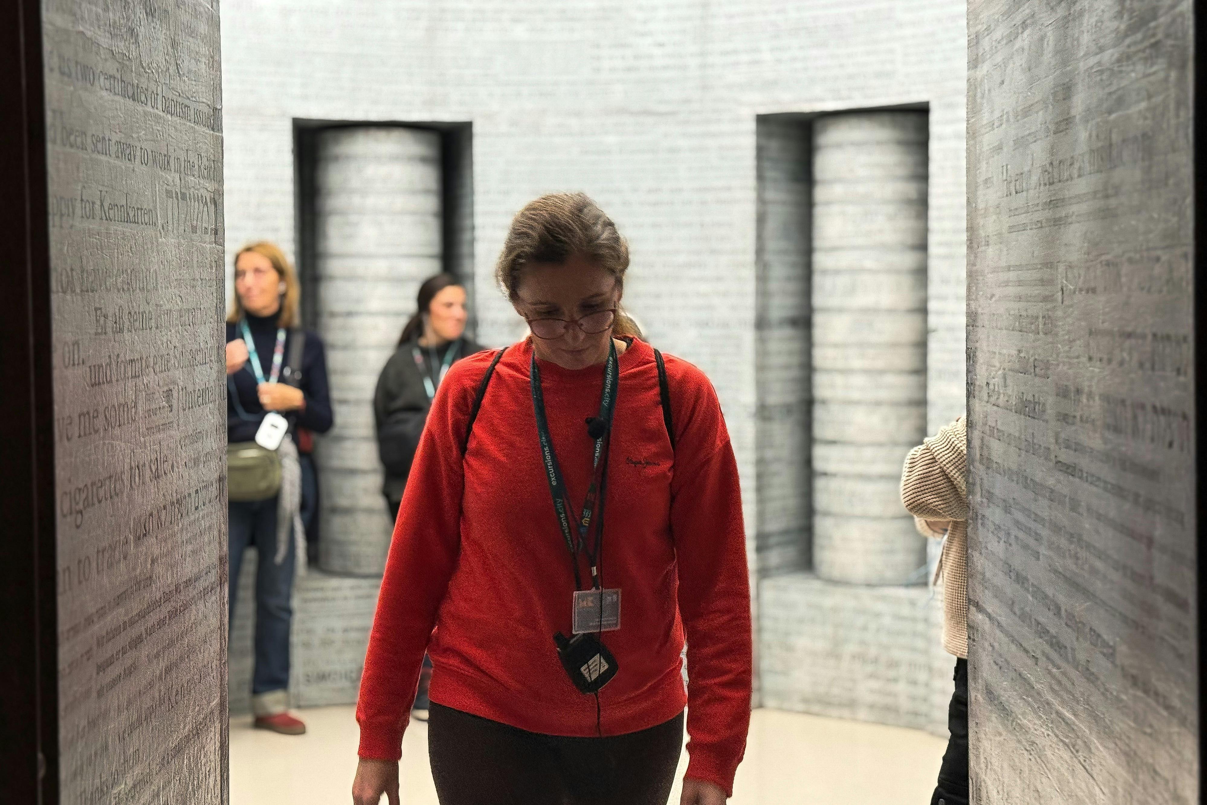 Une femme vêtue d'un pull-over rouge se tient au premier plan, avec deux autres personnes à l'arrière-plan, dans une pièce dont les murs sont recouverts de texte.