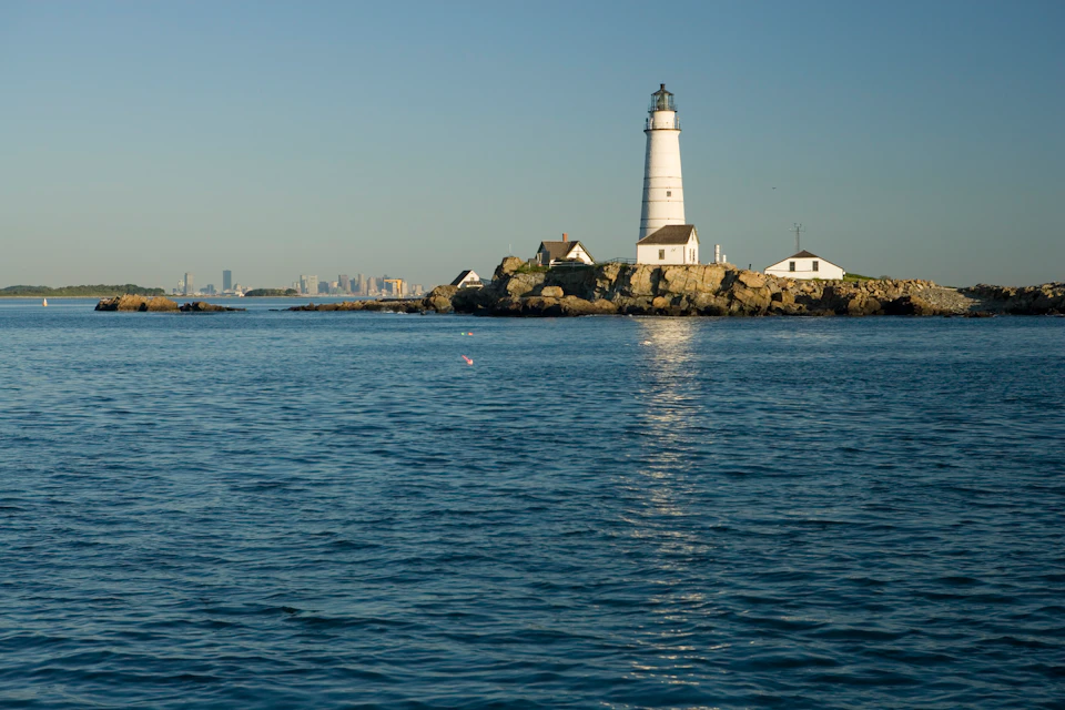 Lighthouse and small buildings on a rocky island, with calm water in the foreground and a city skyline in the distant background.