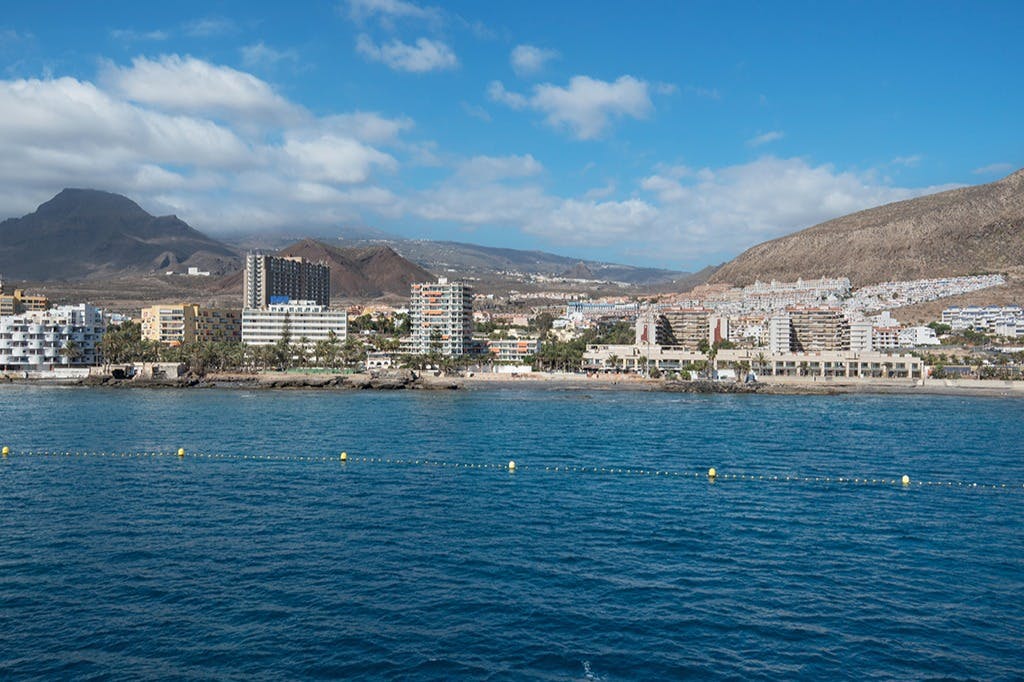Городской пейзаж со зданиями вдоль прибрежной зоны, синим морем на переднем плане и горами на заднем плане под частично облачным небом.