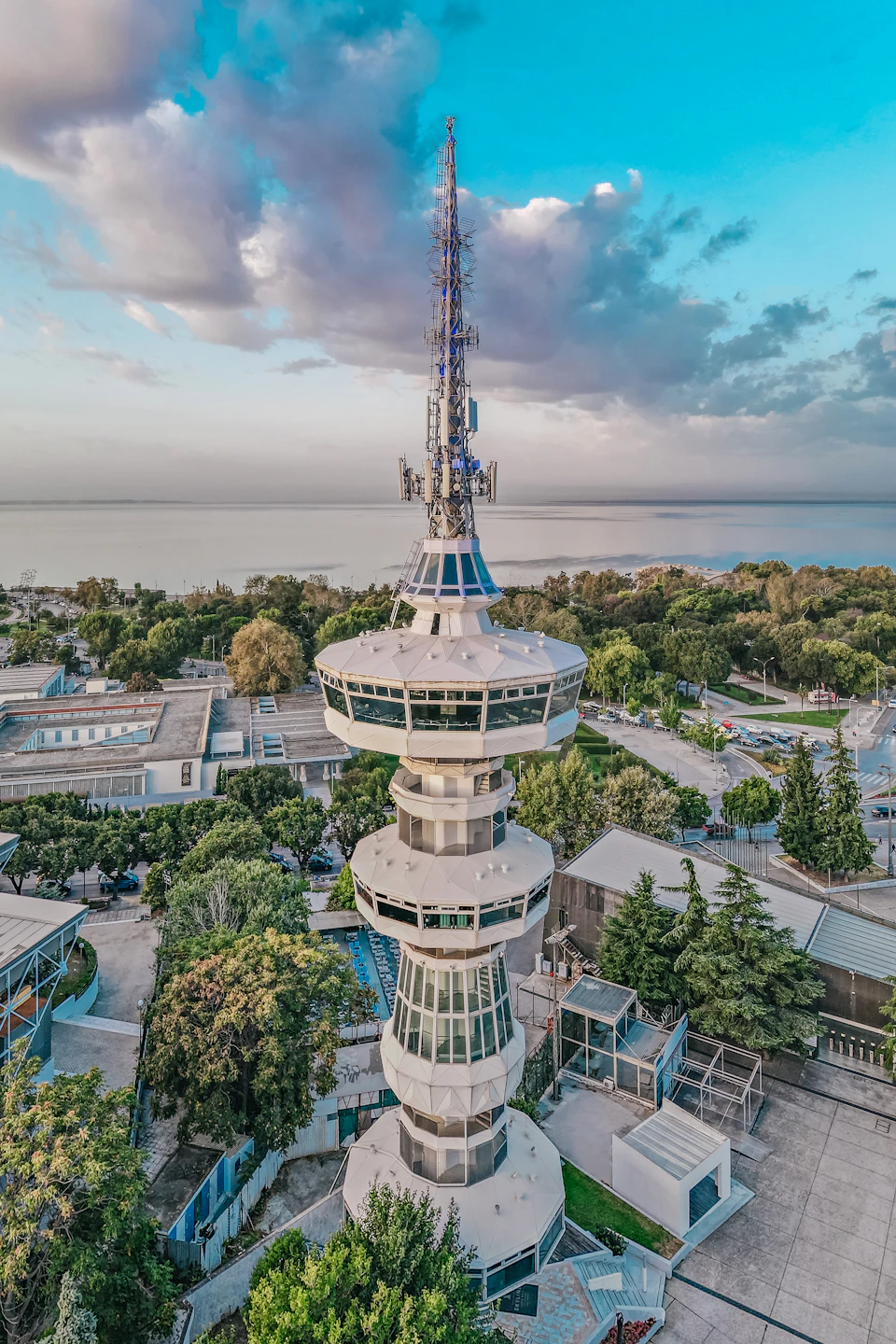 A tall broadcast tower with circular observation platforms, surrounded by trees and buildings near a body of water under a cloudy sky.