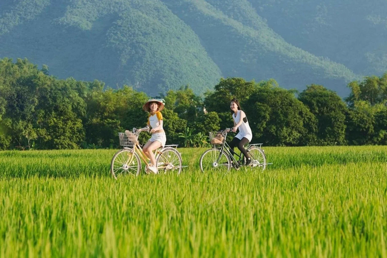 Two women ride bicycles through a lush green field with mountains and trees in the background.