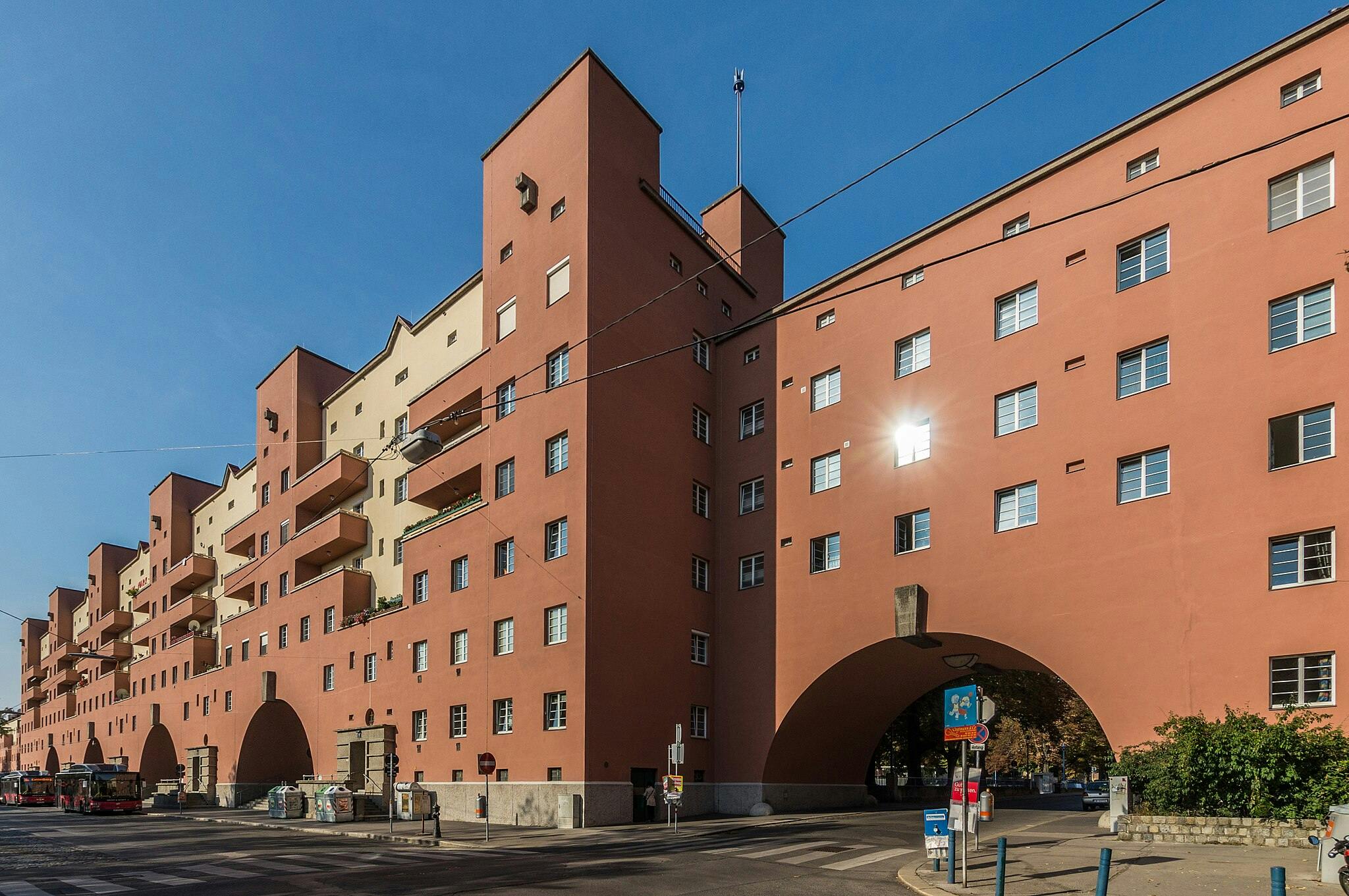 Edifício de apartamentos alto, vermelho, com numerosas janelas e varandas, com grandes arcos na base, contra um céu azul claro.