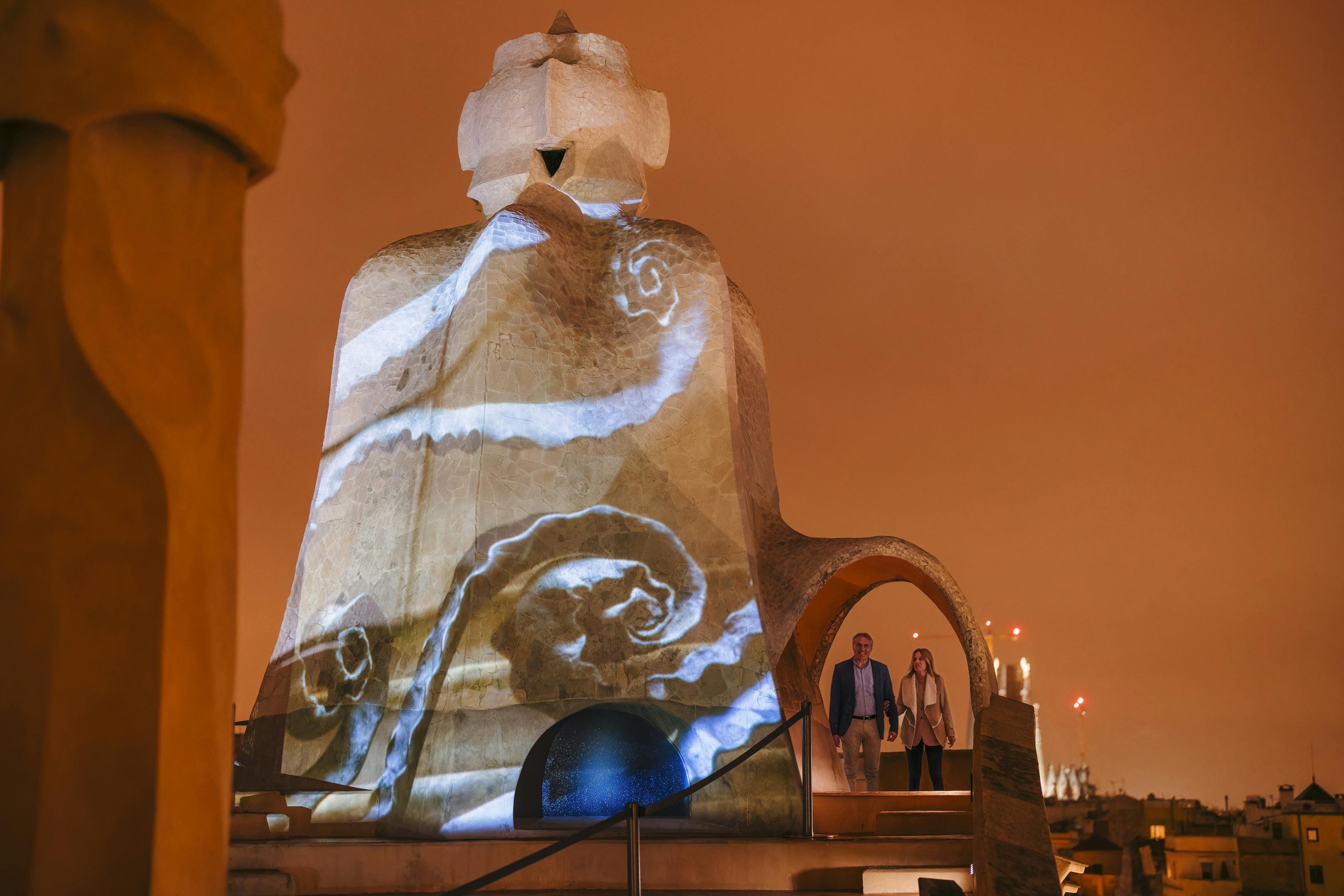 Due persone sono in piedi accanto a una grande scultura in pietra con disegni vorticosi proiettati sotto un cielo arancione e nuvoloso di notte.