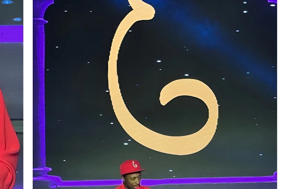 A person in red attire stands on stage with a microphone, in front of a large decorative backdrop, under stage lighting.