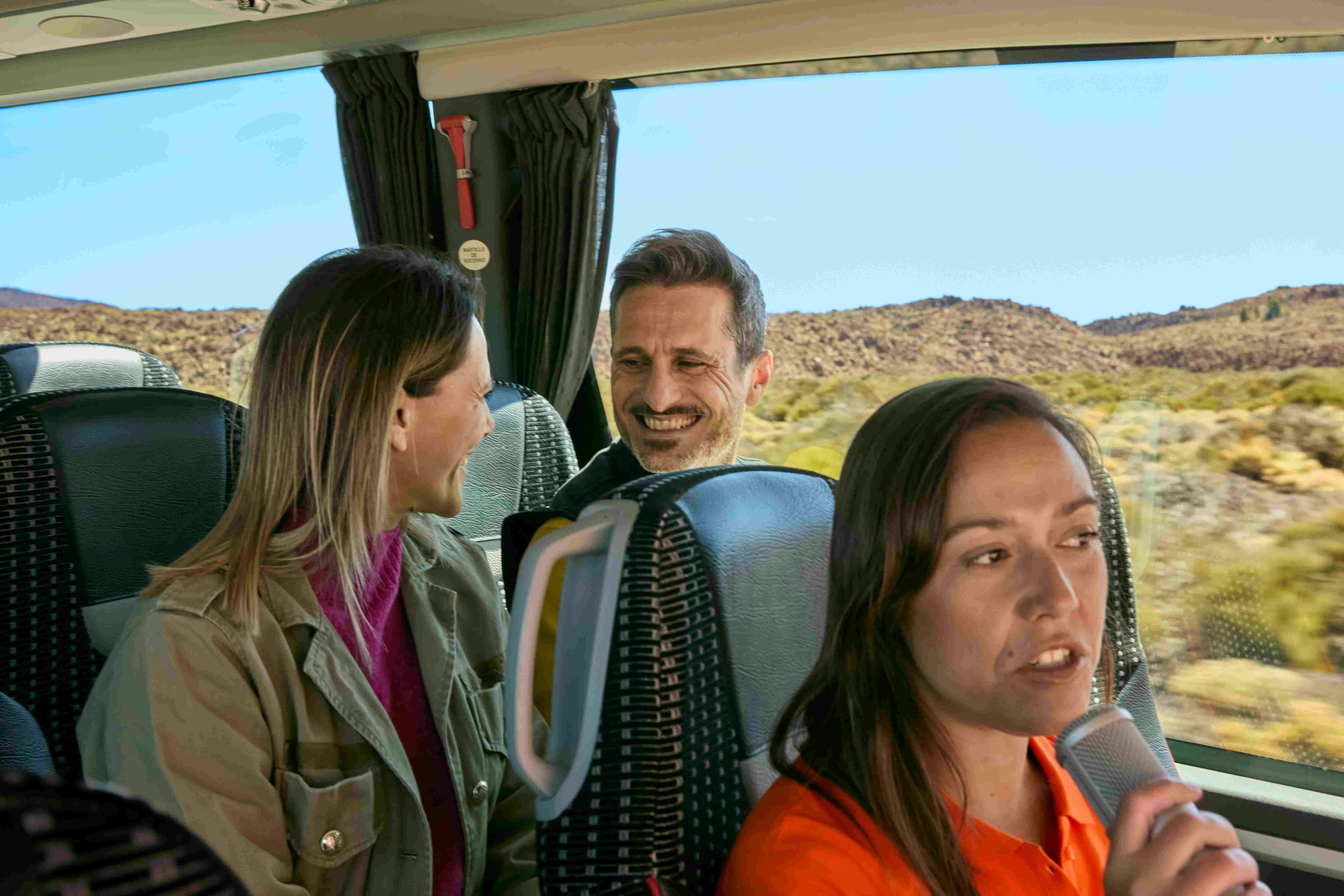 A tour guide speaks into a microphone while two passengers smile on a bus with desert scenery outside the window.