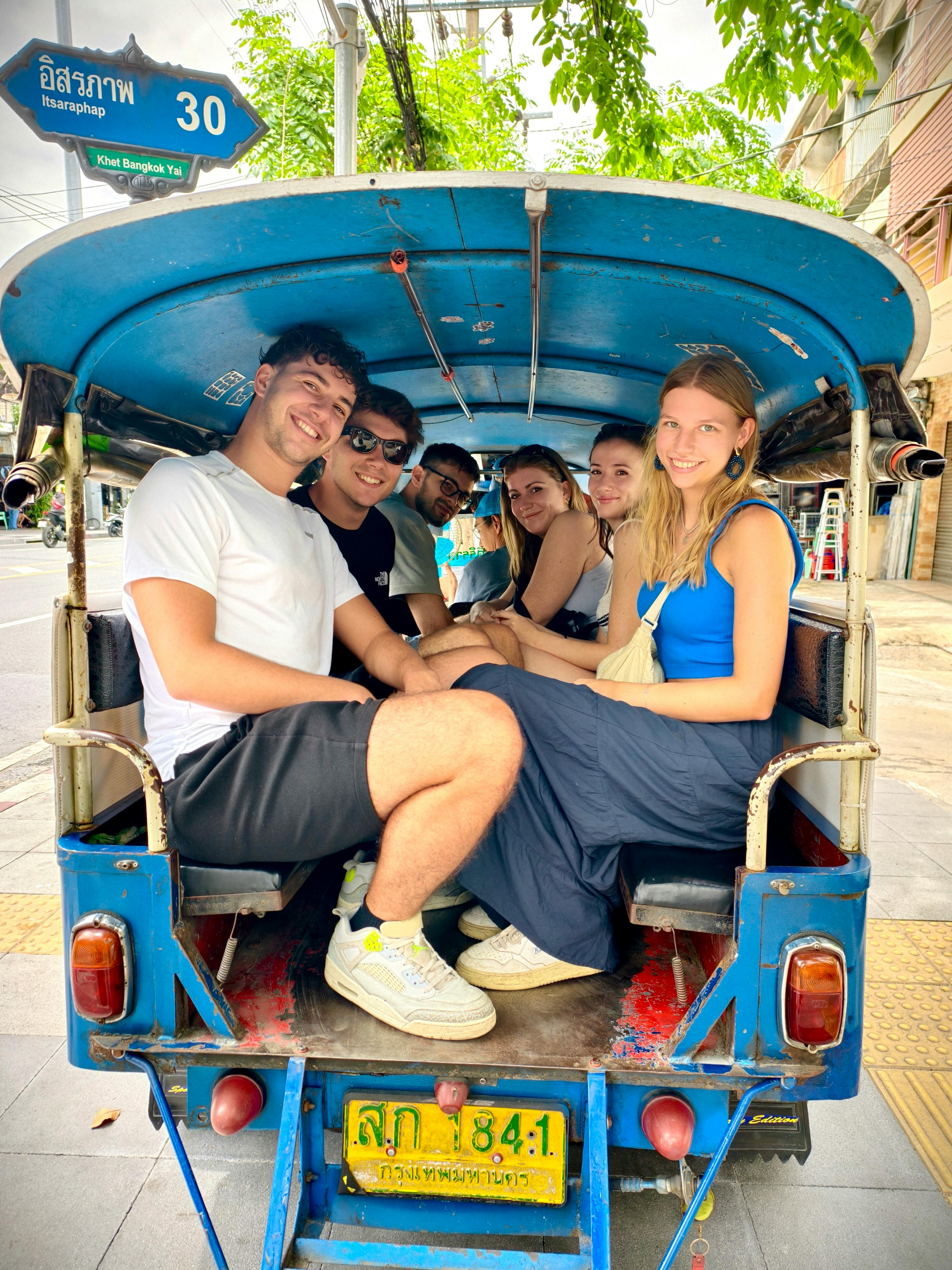 A group of six people sitting together in a tuk-tuk under a blue canopy, parked on a street with buildings and trees in the background.