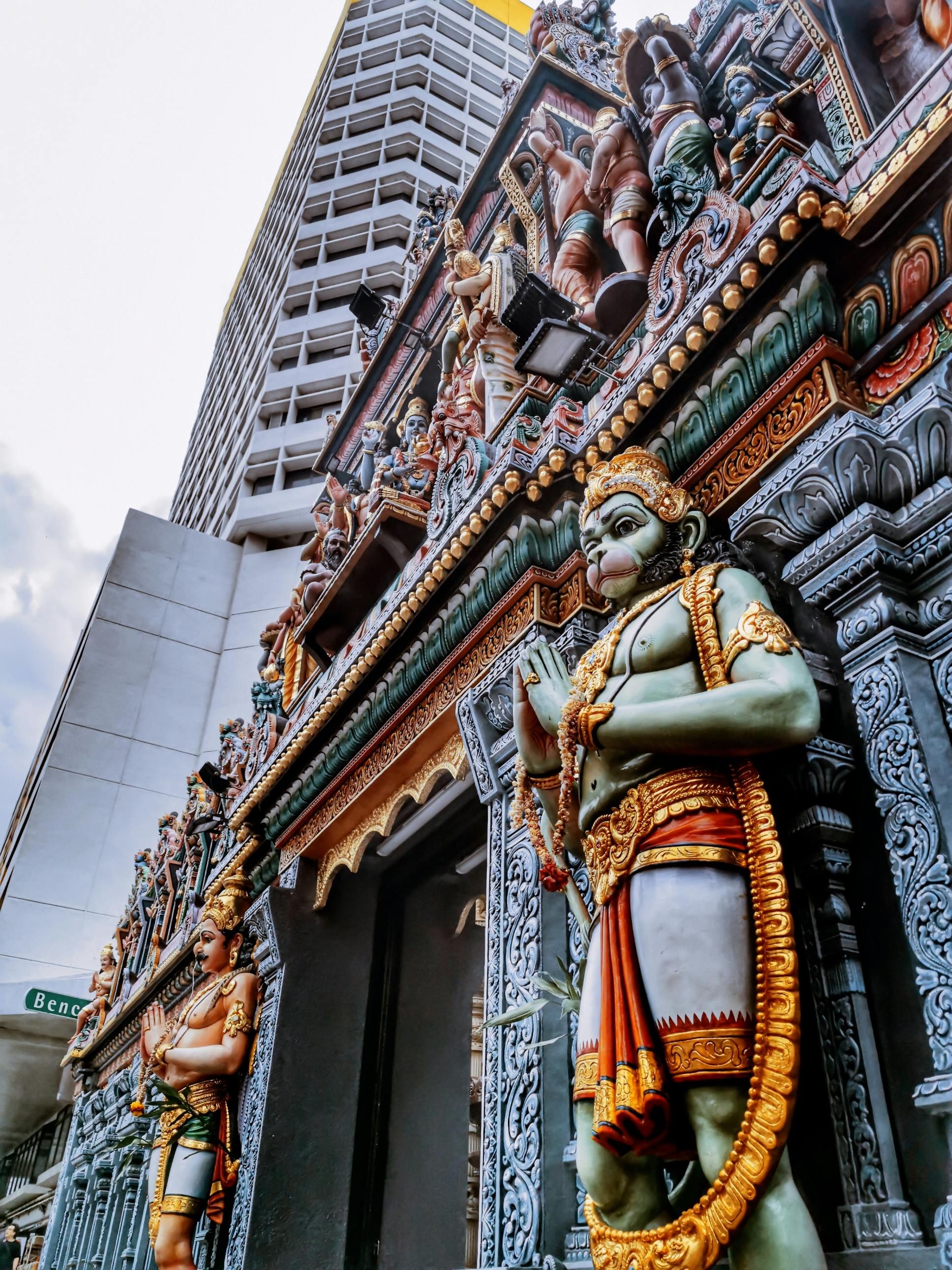 Intricately carved temple facade with colorful statues, set against a modern high-rise building in a cityscape.