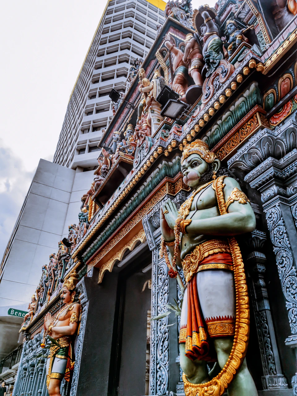 Intricately carved temple facade with colorful statues, set against a modern high-rise building in a cityscape.