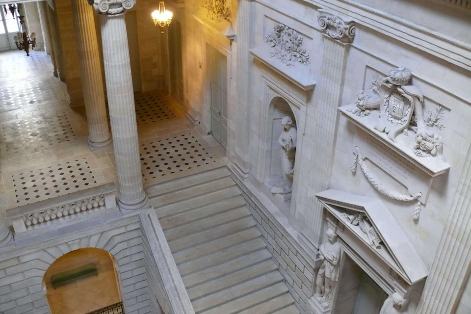 Ornate stone staircase in a grand interior with columns, statues in niches, and detailed wall carvings, lit by a chandelier.