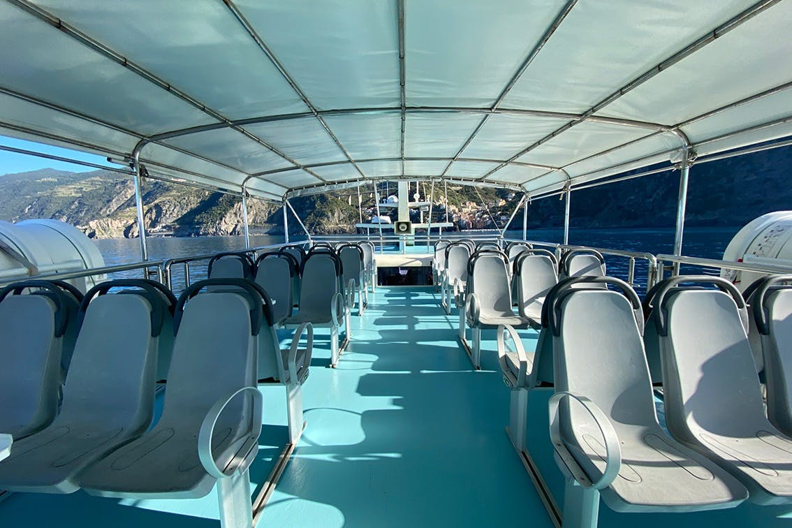 Άποψη ενός άδειου καταστρώματος σκαφών με σειρές γκρίζων καθισμάτων κάτω από ένα θόλο, με θέα τα νερά και τους παράκτιους βράχους στο παρασκήνιο.