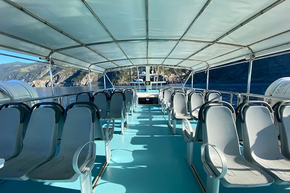 View of an empty boat deck with rows of gray seats under a canopy, overlooking water and coastal cliffs in the background.
