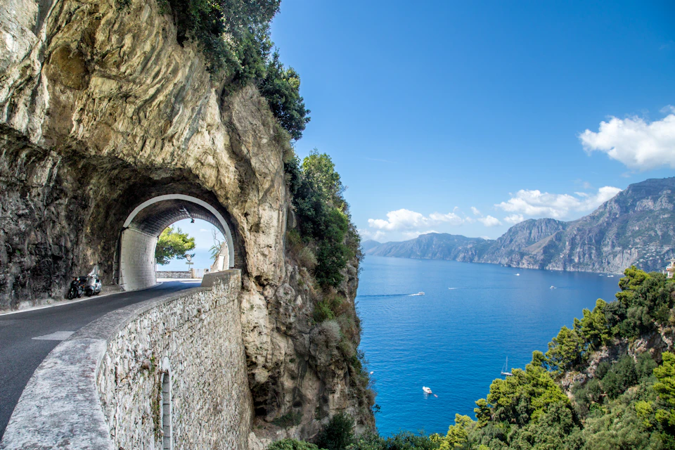 A coastal road passes through a rock tunnel with a view of the sea, cliffs, and boats under a clear blue sky.