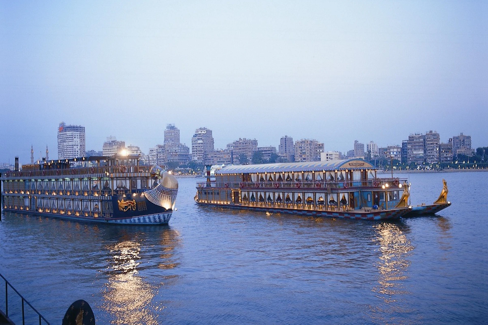 Two large passenger boats on a calm river with a city skyline and several high-rise buildings in the background during twilight.