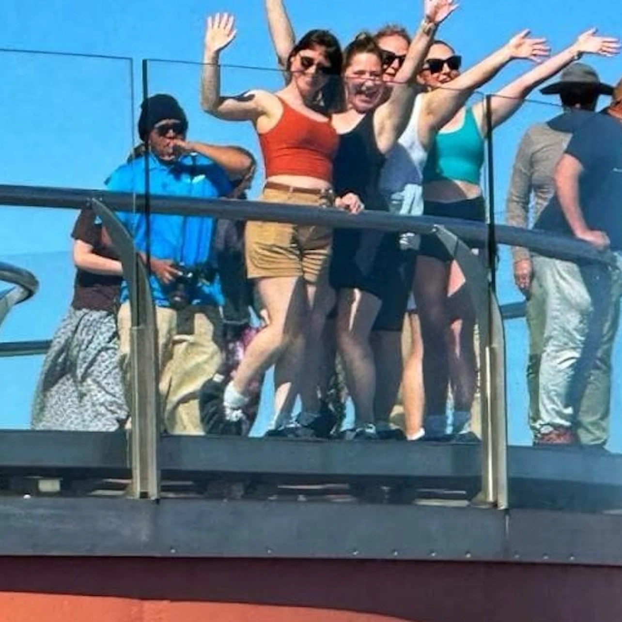 A group of people standing on a glass platform outdoors on a sunny day, some waving and smiling.