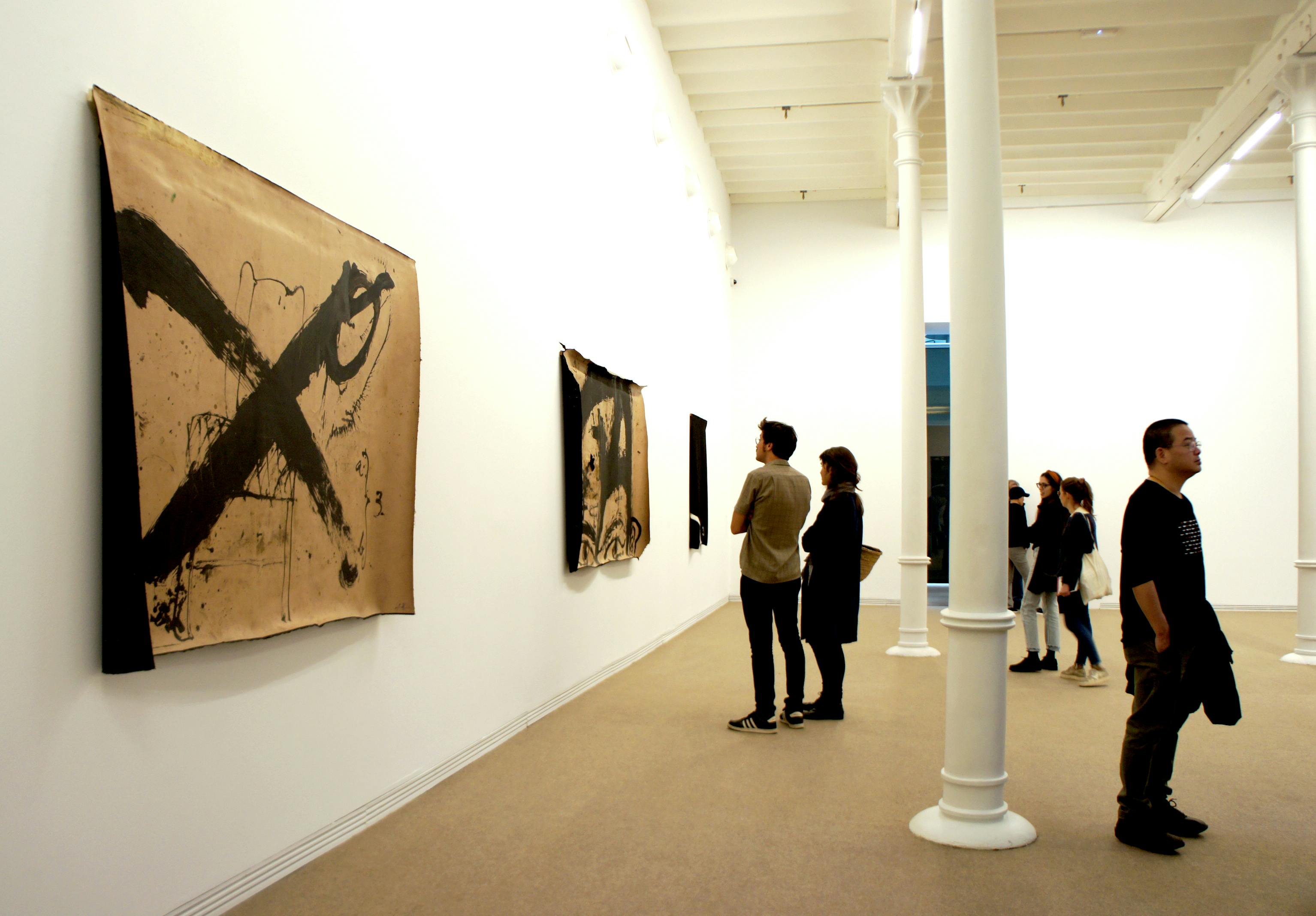 Des personnes observent des œuvres d'art sur des murs blancs dans une galerie bien éclairée avec des colonnes blanches.