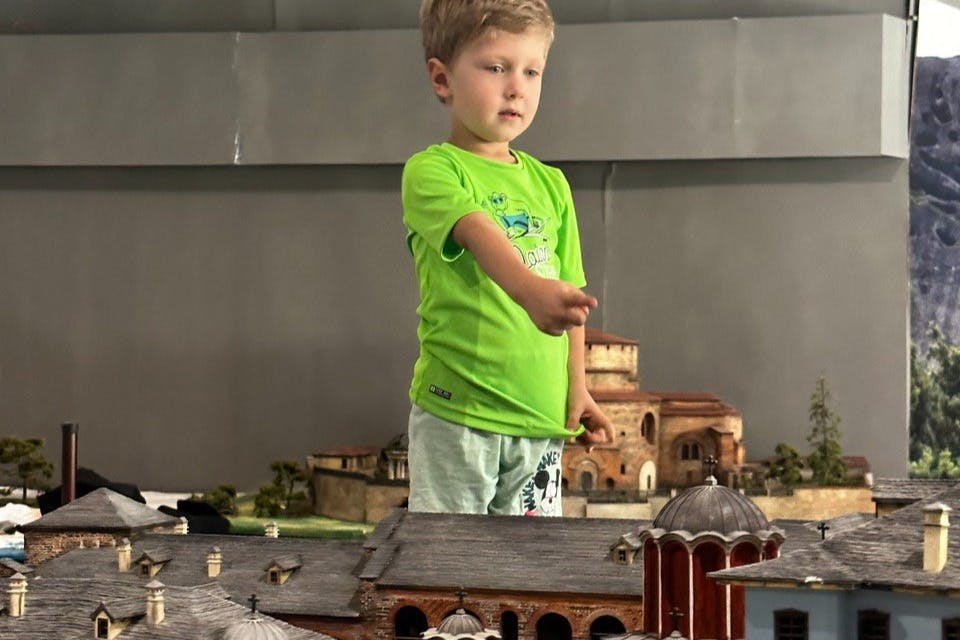 A young boy in a green shirt stands near a detailed architectural model of buildings with domes and arches.