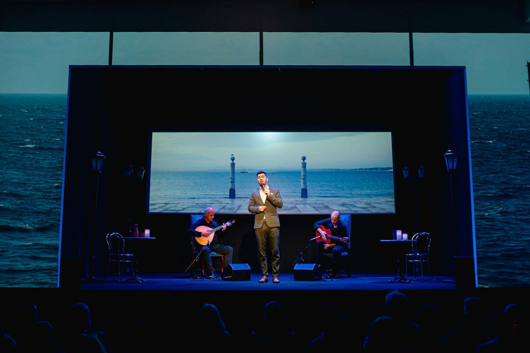 2人のギタリストが両側に座った状態で、歌手がステージ上で演奏します。 背後にある大画面には、海辺の光景が映し出されている。