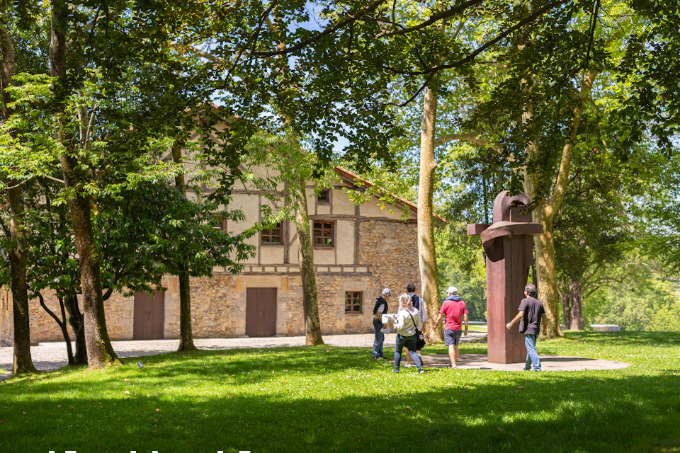 People walking through a grassy park with trees, near a stone building and a large metal sculpture.