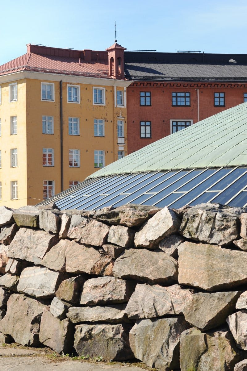 Stone wall in the foreground, sloped glass roof behind it, and yellow and red brick buildings in the background.