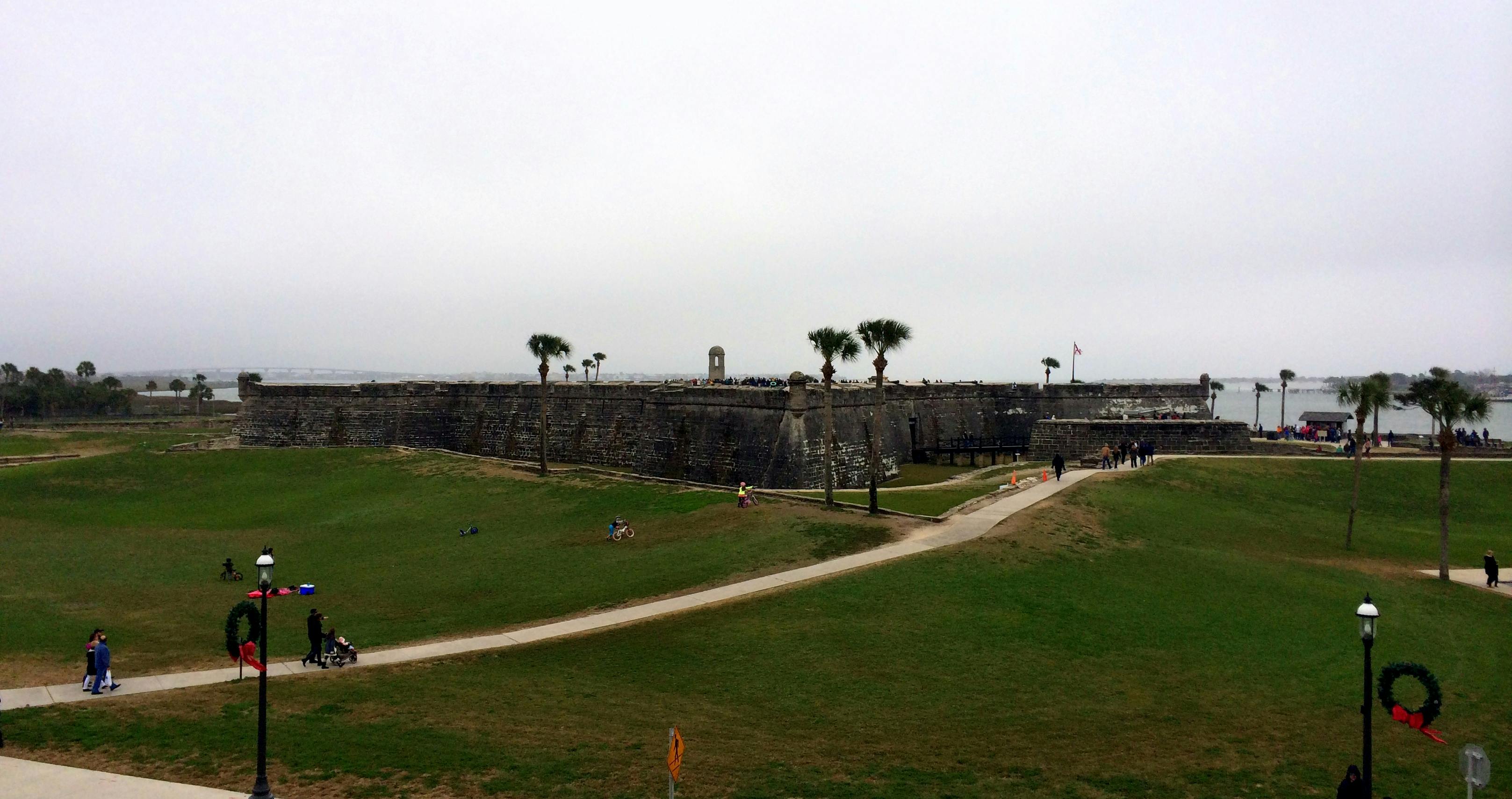 石头堡垒，草地上有棕榈树，有几个人散步，还有多云的天空。 通往入口的通道。