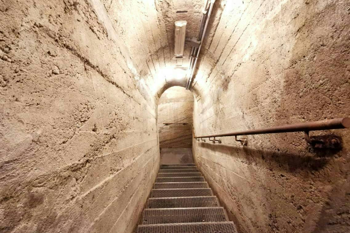 Una estrecha escalera de hormigón con peldaños y barandillas metálicas, iluminada por luces de techo, que baja hacia abajo.