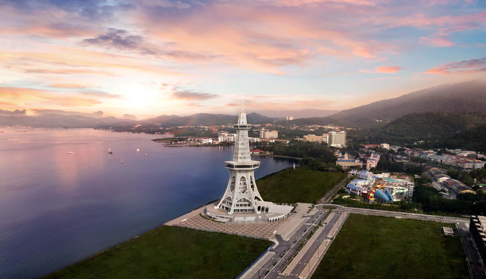 A modern tower near a waterfront, surrounded by green space and roads, with a cityscape and hills in the background.