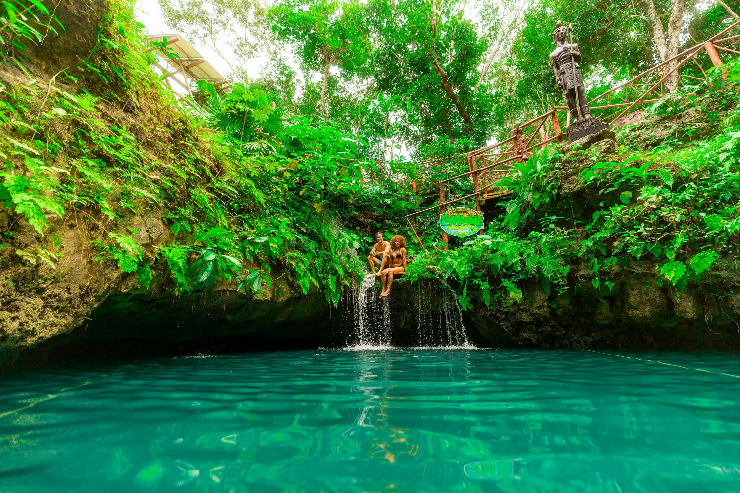 Dues persones s'asseuen en un petit penya-segat damunt d'una cascada envoltada d'una vegetació exuberant, amb una piscina d'aigua blava a sota.
