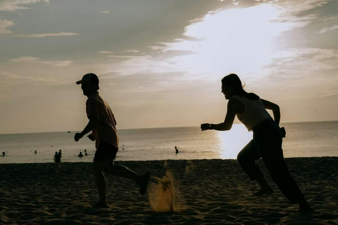 Silhouettes of two people running on a sandy beach at sunset, with the ocean and a bright sky in the background.