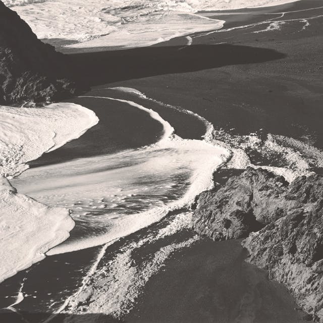 波浪在崎岖的海岸线上的深色沙滩上起泡，前景是岩石和阴影。