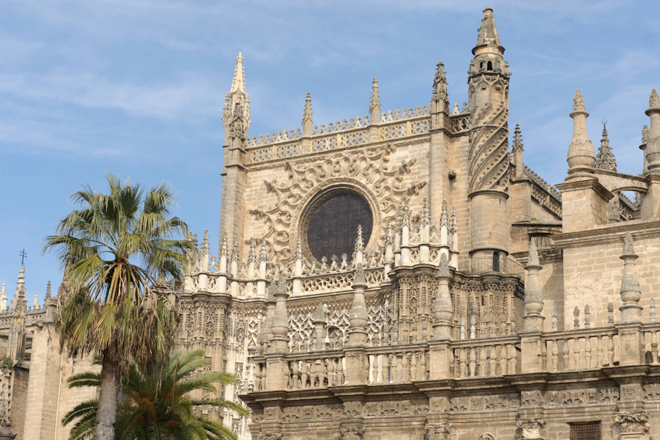 Gothic cathedral facade with intricate stone carvings, a large circular window, and palm trees in the foreground.