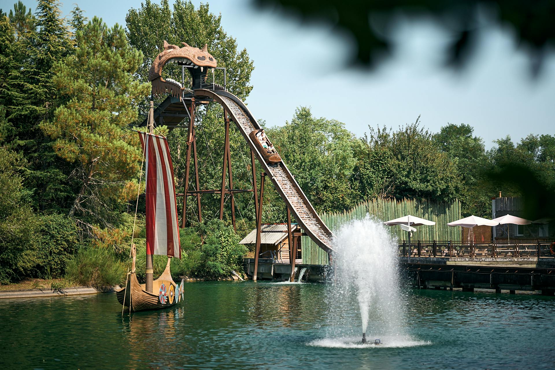 Jízda na vodě se strmým pádem do bazénu, vikingská loď s plachtou a fontána v popředí obklopená zelení.