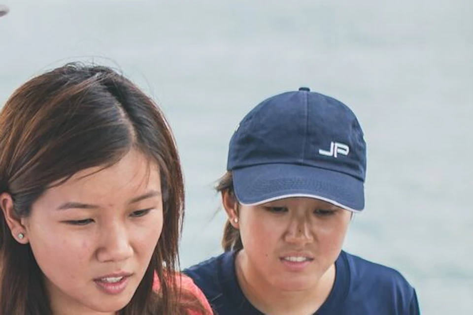 Four people on a boat, one person steering while others observe. One person wearing a hat points towards the boat's controls.