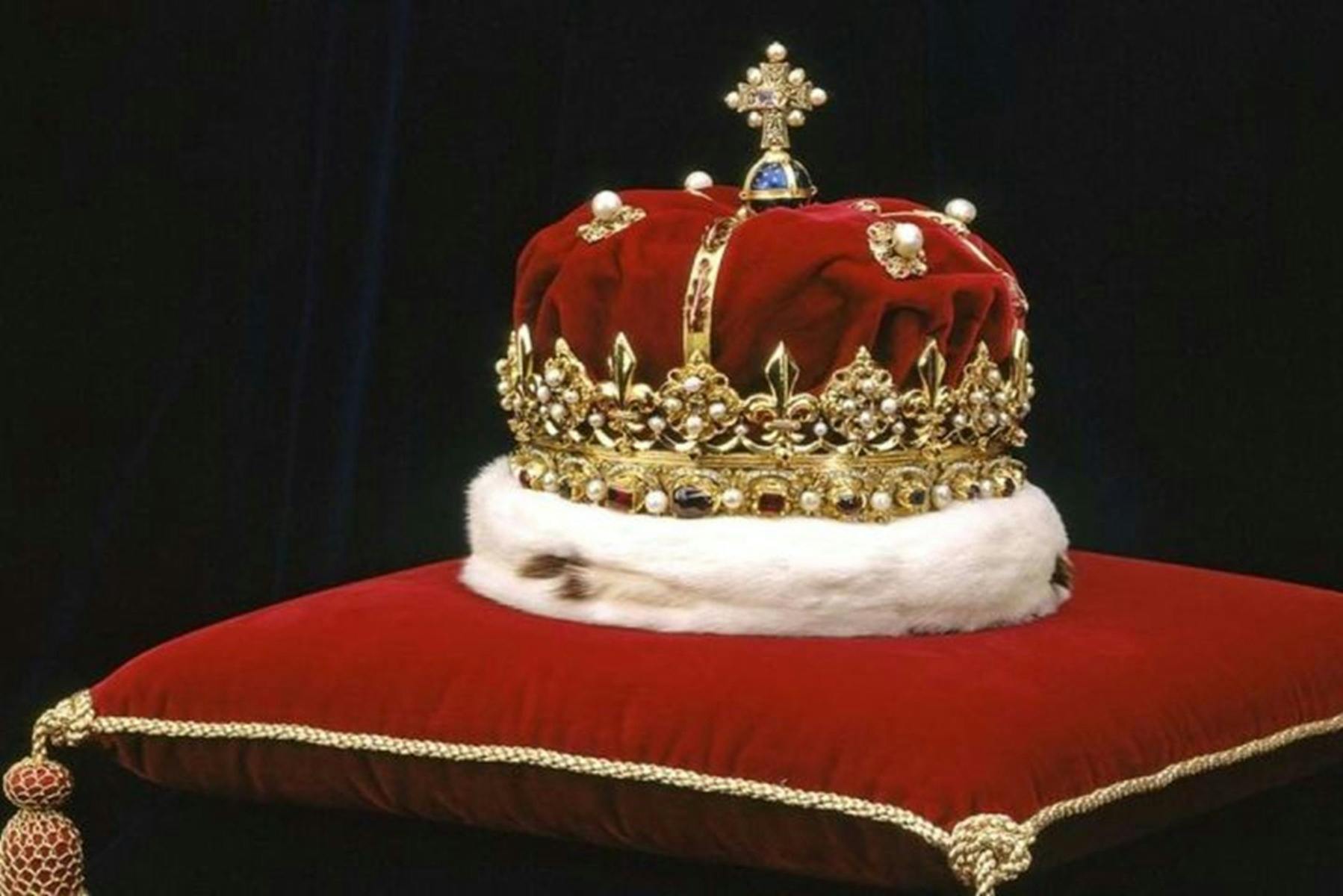 Una corona regale ornata di gioielli e oro, appoggiata su un cuscino di velluto rosso con finiture dorate su uno sfondo scuro.