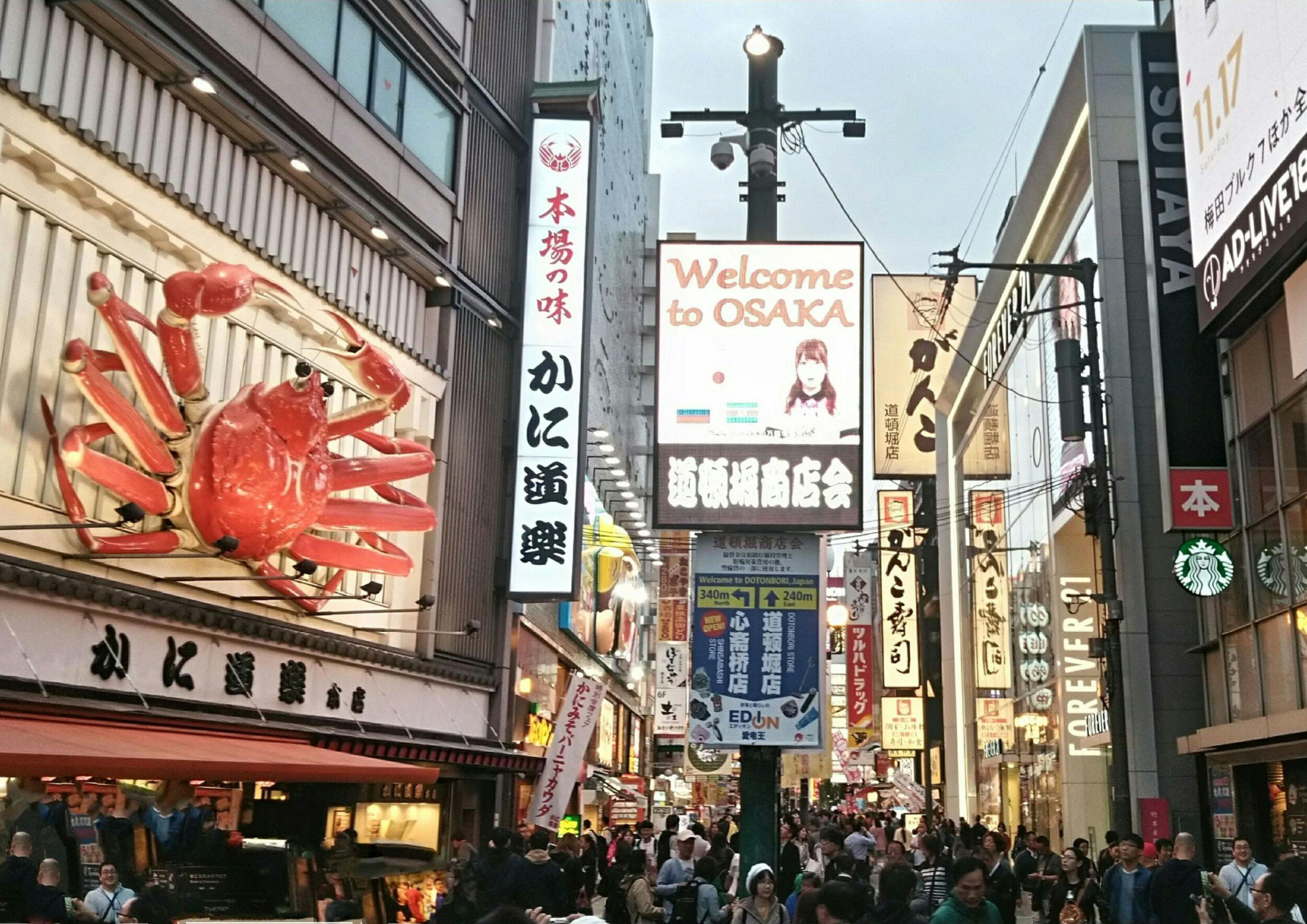A busy street in Osaka with colorful signs, a giant crab model, and a crowd of people walking under a cloudy sky.