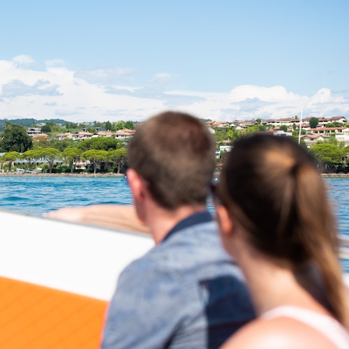 Two people facing away, riding a boat on a lake, with houses and trees visible in the background under a clear blue sky.