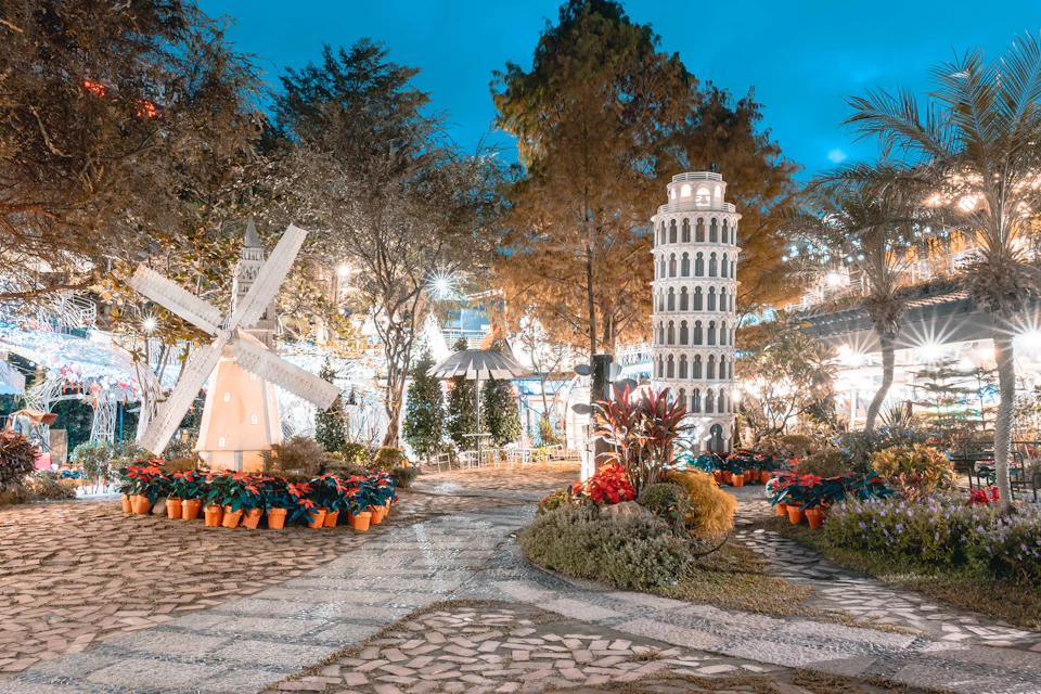A garden scene with a windmill and a replica of the Leaning Tower of Pisa, surrounded by plants and illuminated by lights.